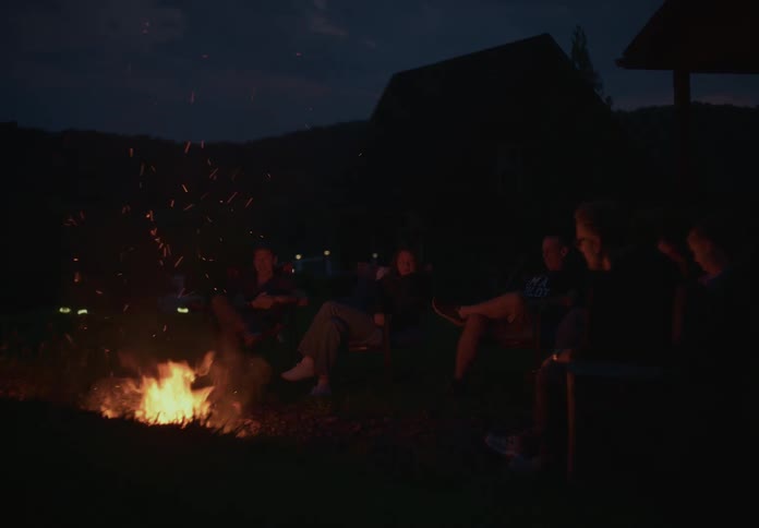 A group of friends sit around a crackling campfire in the backyard on a peaceful evening, talking and relaxing