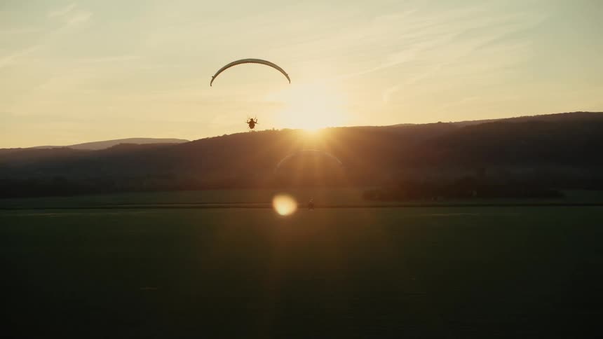Two paragliders gracefully soar through the sky, silhouetted against a stunning golden sunset over a vast rural landscape
