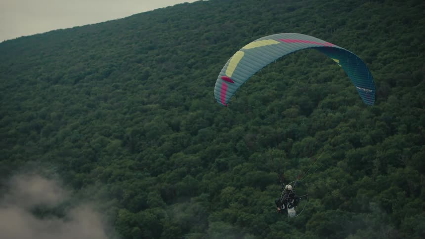 A powered paraglider soars gracefully over a vast green forest on a misty, overcast day