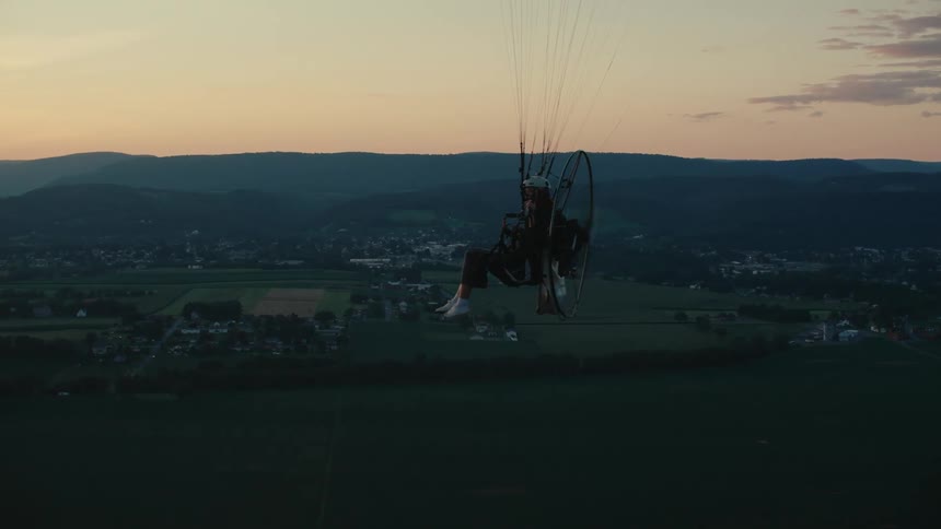 An aerial view of a person flying a powered paraglider over a vast rural landscape during a serene sunset