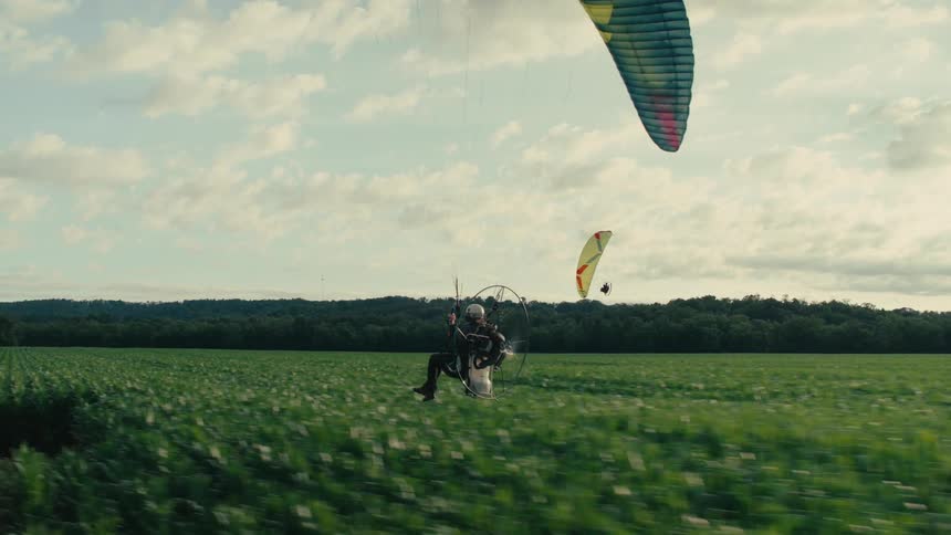 A tracking shot follows a powered paraglider as it soars low over a lush green cornfield on a partly cloudy day
