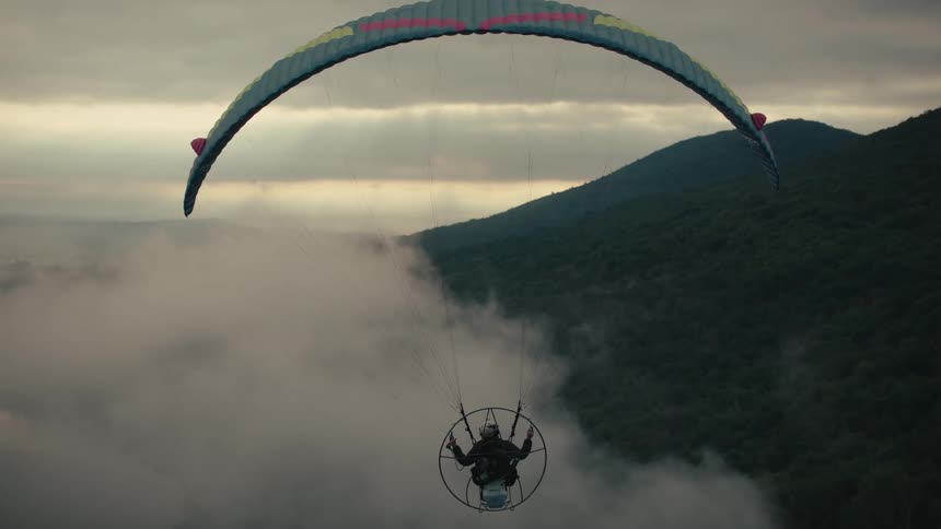 Cinemagraph of a powered paraglider hovering over a sea of clouds near a forested mountain at dusk