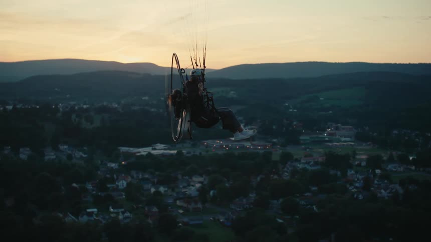 Cinematic slow-motion view of a paramotor pilot flying high above a quiet town surrounded by mountains at sunset