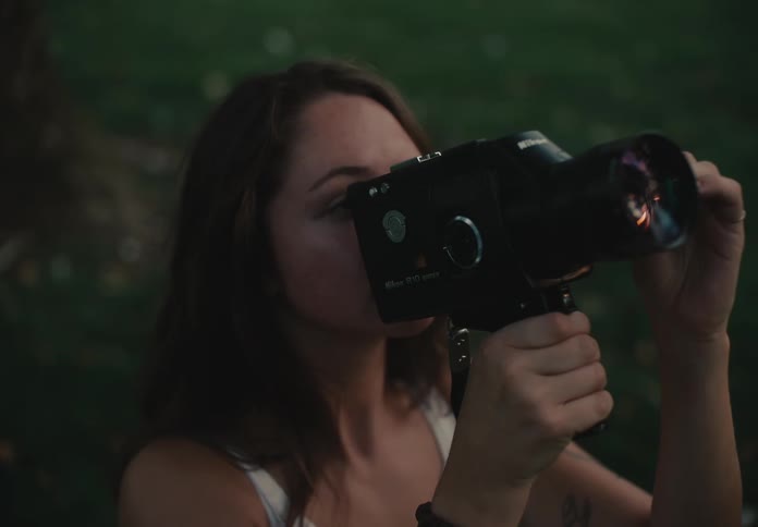 A thoughtful young woman operates a vintage Super 8 film camera while sitting in a park at dusk