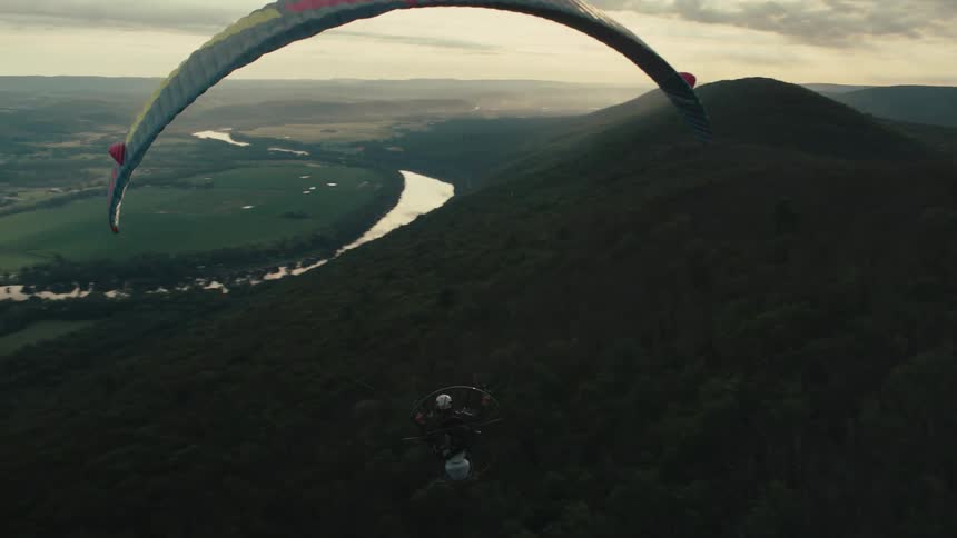 An epic aerial shot follows a powered paraglider soaring over a lush, forested mountain valley and winding river at golden hour