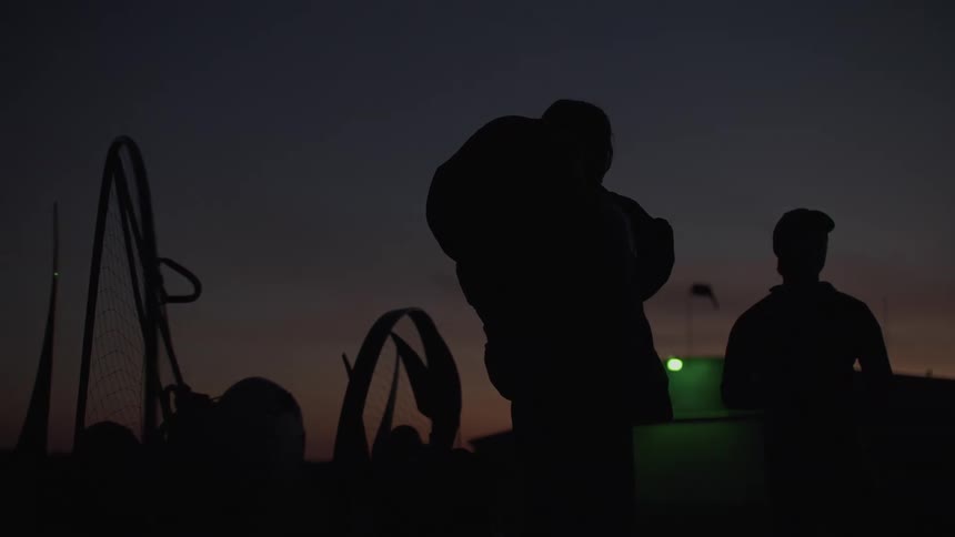 Silhouettes of paramotor pilots prepare their gear as the camera slowly pans across an airfield at dusk