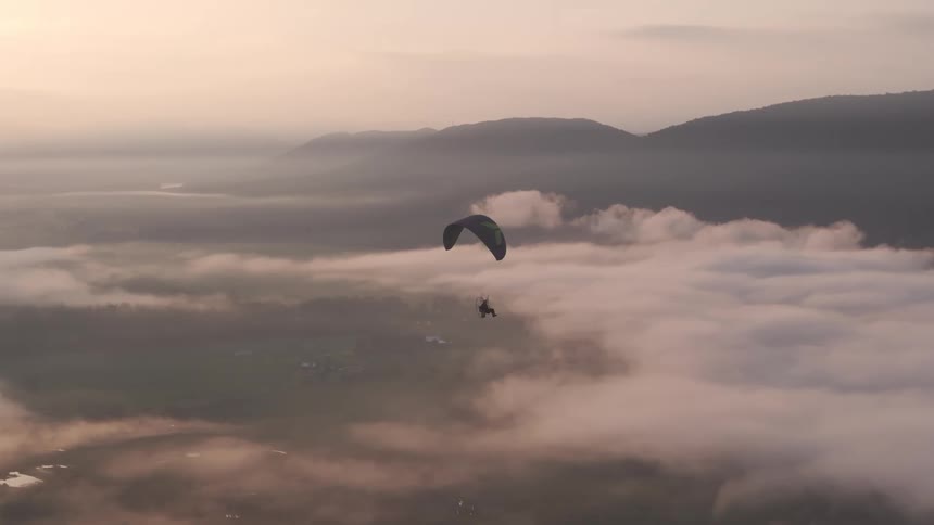 A powered paraglider soars majestically above a sea of clouds in a mountain valley during a serene sunrise