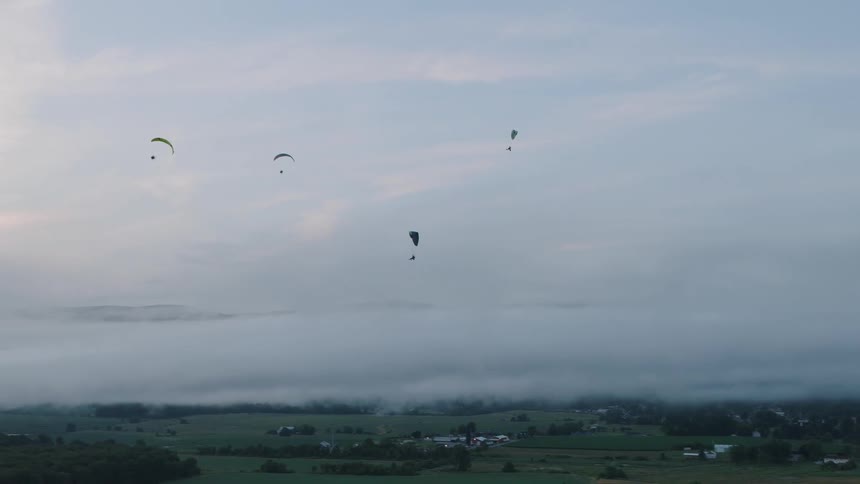 A group of paragliders soar high in the sky above a foggy rural valley at dawn