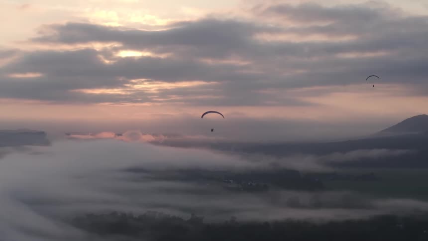 A breathtaking aerial view shows paragliders peacefully soaring above a thick blanket of fog at sunrise