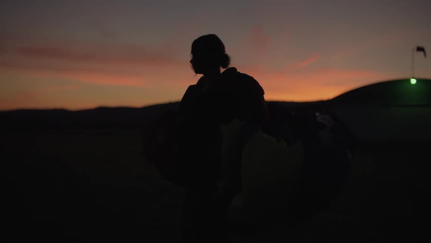 A female skydiver in slow motion gathers her parachute in silhouette against a vibrant sunset sky
