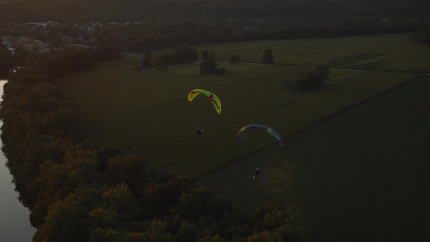 Aerial view of three powered paragliders flying together over a rural landscape of fields and a river during a tranquil golden hour sunset
