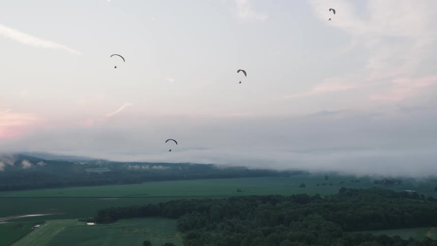 A group of paragliders fly serenely above a sea of clouds over a lush green landscape during a beautiful sunrise