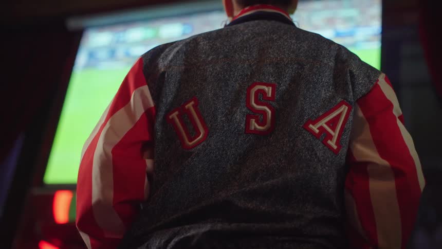 A patriotic fan wearing a USA jacket intently watches a live sports game on a large screen in a dimly lit room
