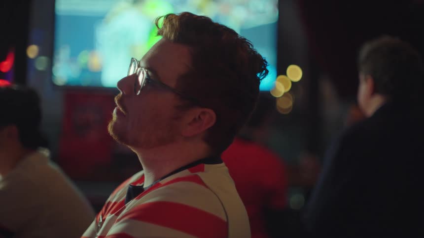 A pensive young man with red hair and glasses is captivated by a sports game on TV in a dark bar