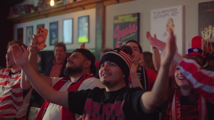 Enthusiastic sports fans wearing team colors clap and cheer together while watching a game in a lively bar