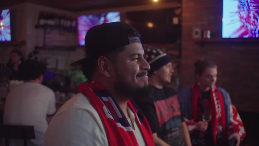 A happy man wearing a team scarf smiles while watching a soccer game with friends at a sports bar
