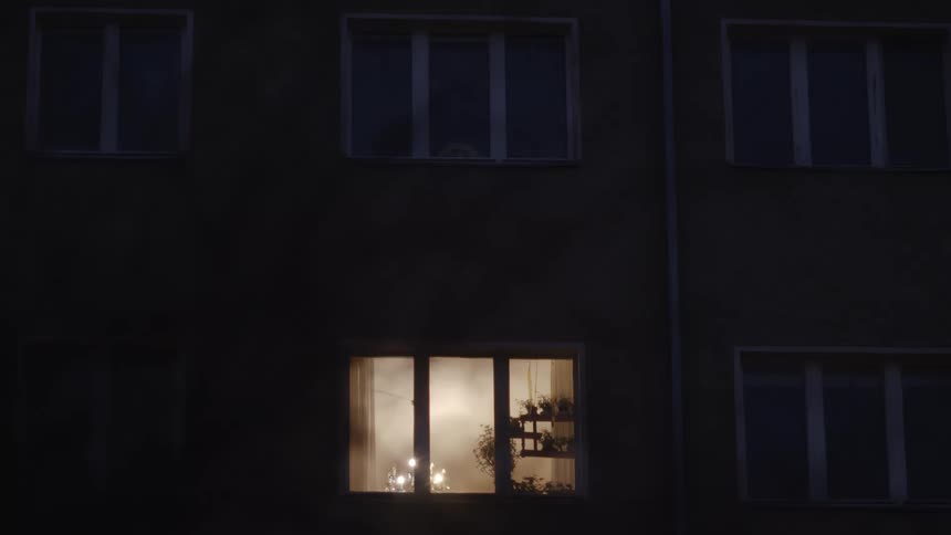 A hazy, warm light glows from a single apartment window on a dark night, surrounded by the unlit windows of the building