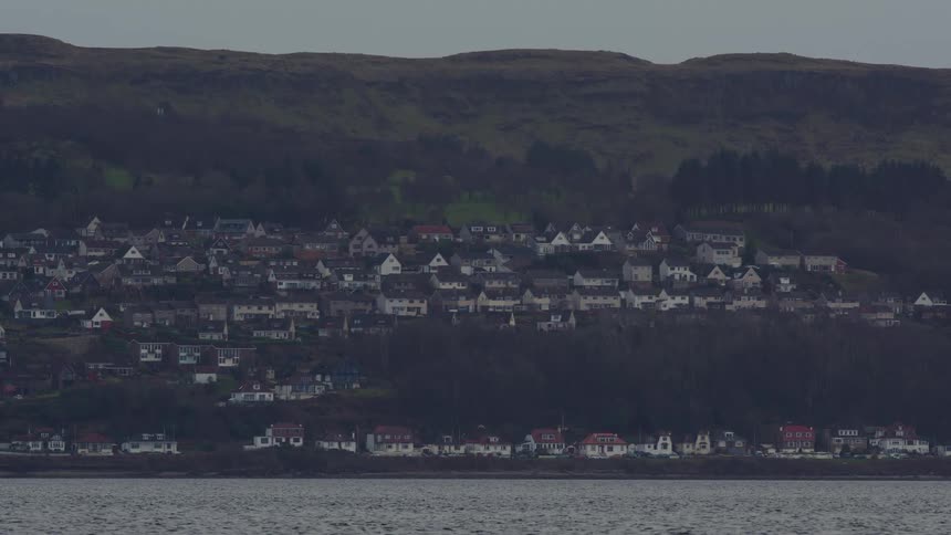 A tranquil view of a seaside village nestled at the base of a large, green hill across the water