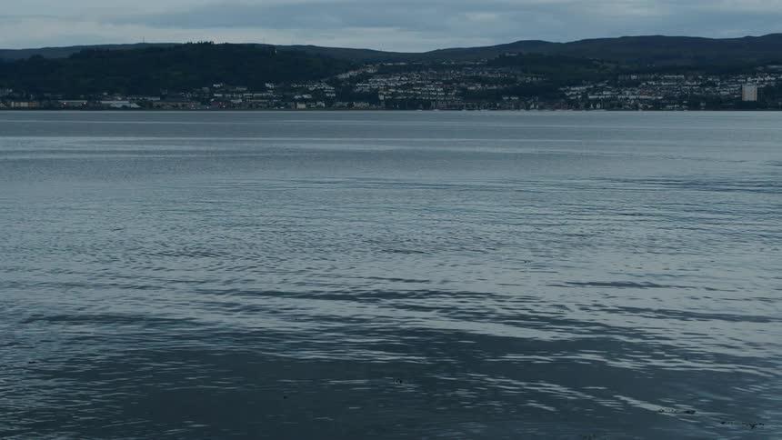 A serene view of a calm loch with a distant town on the hilly shoreline under an overcast sky