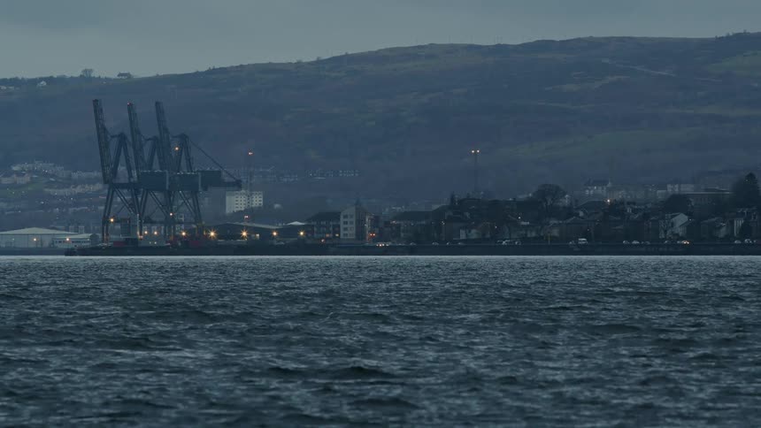 A wide view across a choppy sea shows an industrial port with large cranes and a city at the base of hills on an overcast day