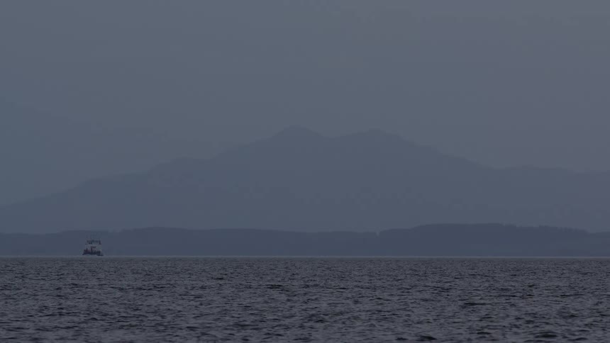 A single boat floats on a vast ocean with a foggy mountain range silhouetted against a grey, overcast sky