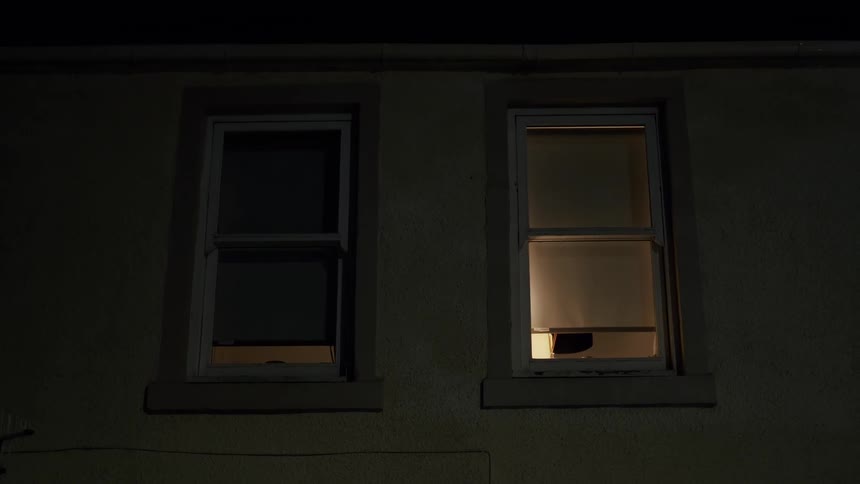 A static shot shows two windows of a house at night, with one room illuminated from within