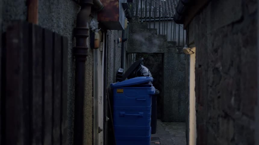 Steam rises from a pipe in a dark, grimy urban alleyway next to an overflowing blue recycling bin