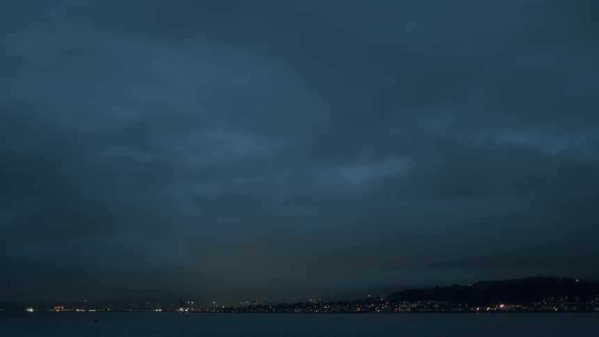 A dramatic wide shot of a distant cityscape twinkling at night under a dark, cloudy sky over the water