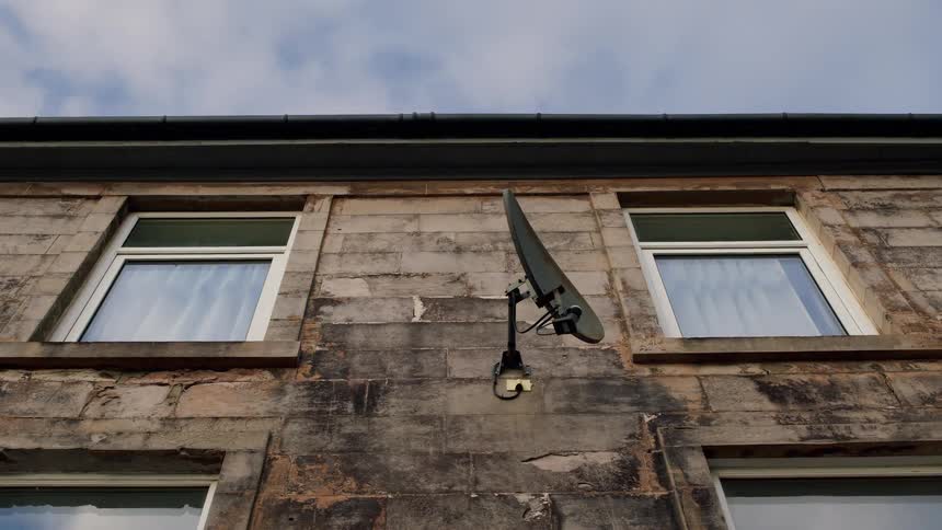Hand-held shot of a satellite dish mounted on the exterior wall of an old stone building