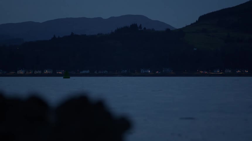 A calm loch at dusk with a flashing green navigation buoy and the lights of a distant village nestled at the foot of mountains