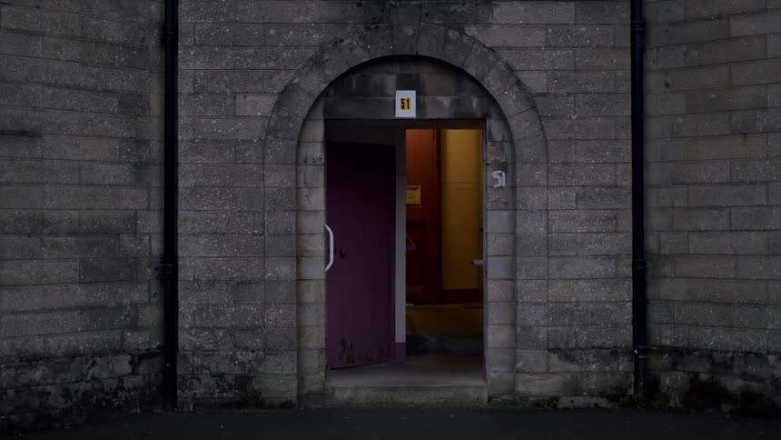 A static shot shows a dark, arched stone doorway with the inner hallway illuminated at dusk