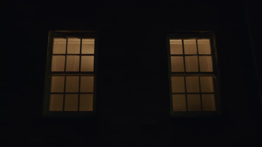 Two windows with closed blinds glow with warm light against the pitch-black exterior of a building at night
