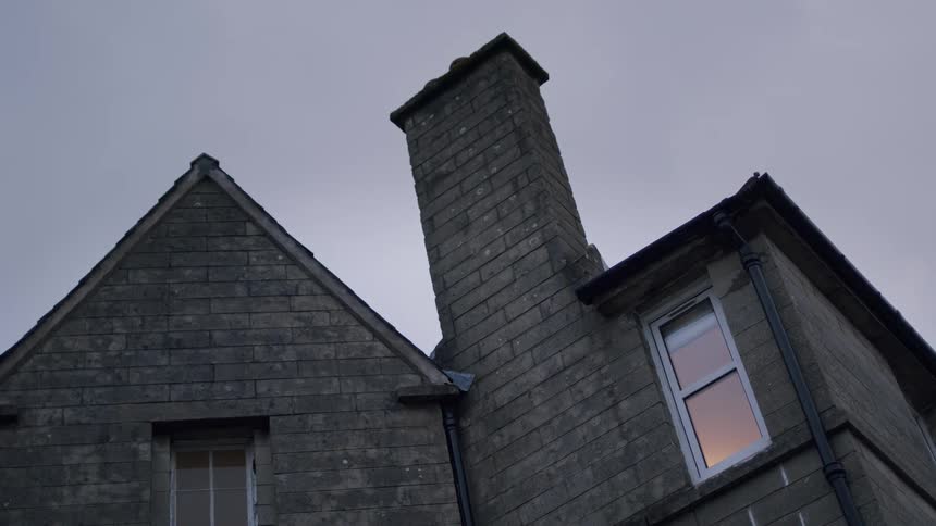 A static low-angle shot captures an old stone house at dusk, with the warm colors of sunset reflected in a window