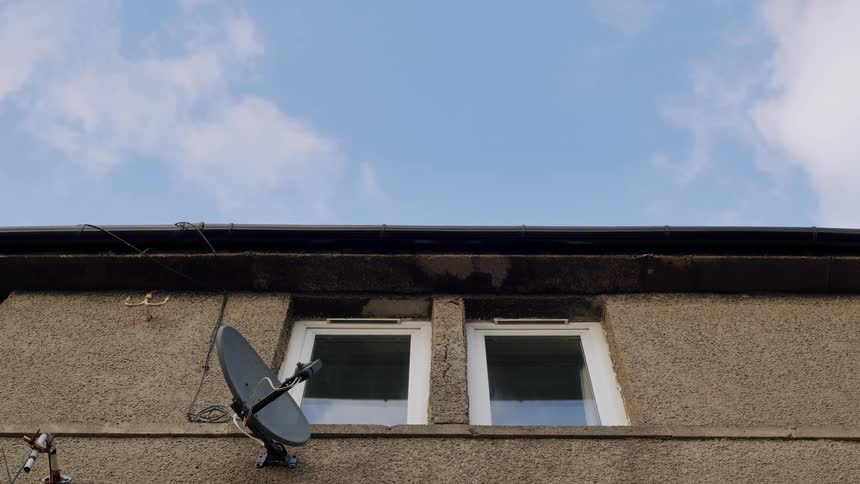 Blue sky and clouds drift over the top windows of a UK terraced house with a satellite dish