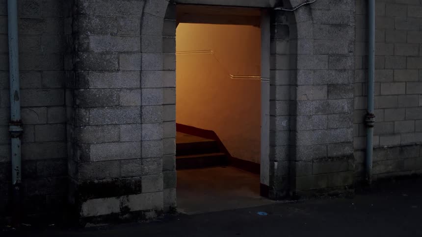 A mysterious open doorway in a dark stone wall at night reveals a brightly lit staircase leading downwards