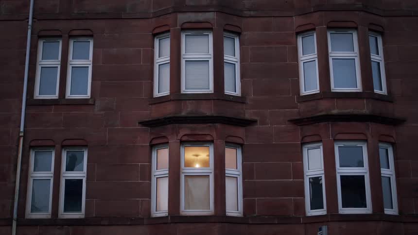 A single illuminated window glows warmly on the dark facade of a red stone apartment building at dusk