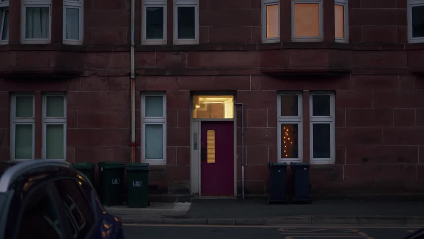 A handheld exterior shot of a red stone tenement building at dusk, where the entrance and windows are warmly lit