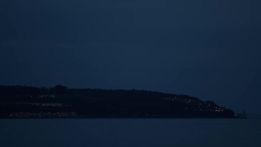 Twinkling lights from a distant coastal town dot the silhouetted shoreline under a dark blue night sky