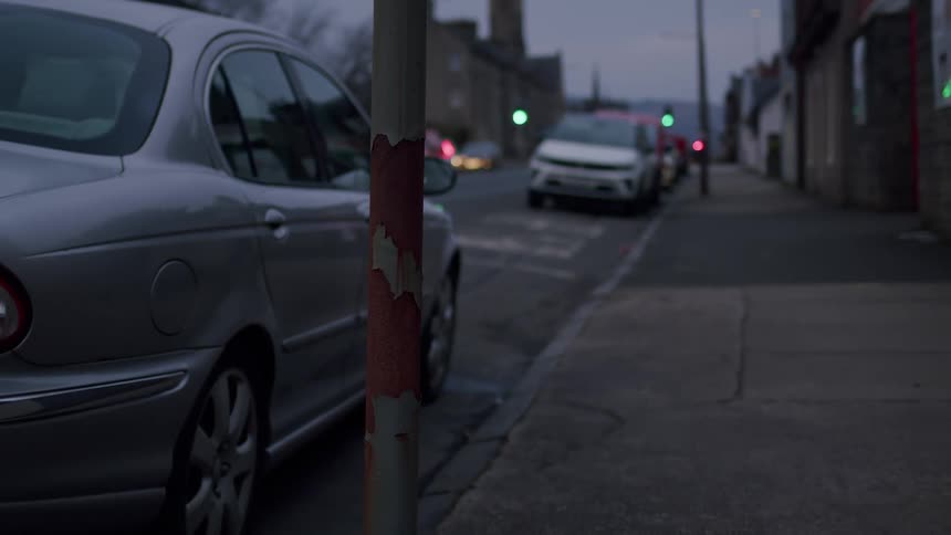 A rusty pole stands on a quiet urban sidewalk at dusk, with parked cars and distant traffic lights in the background