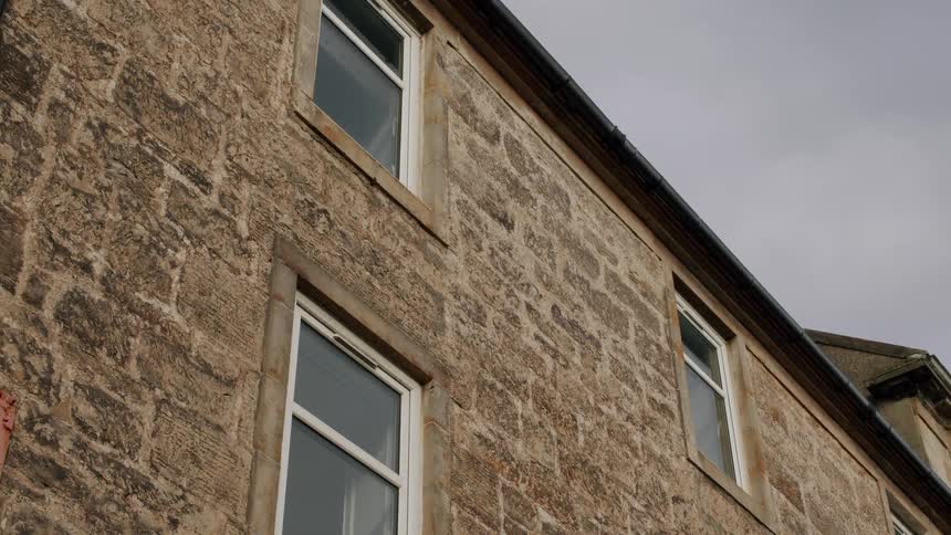 A static, low-angle shot of an old stone building facade with white-framed windows against a grey, overcast sky