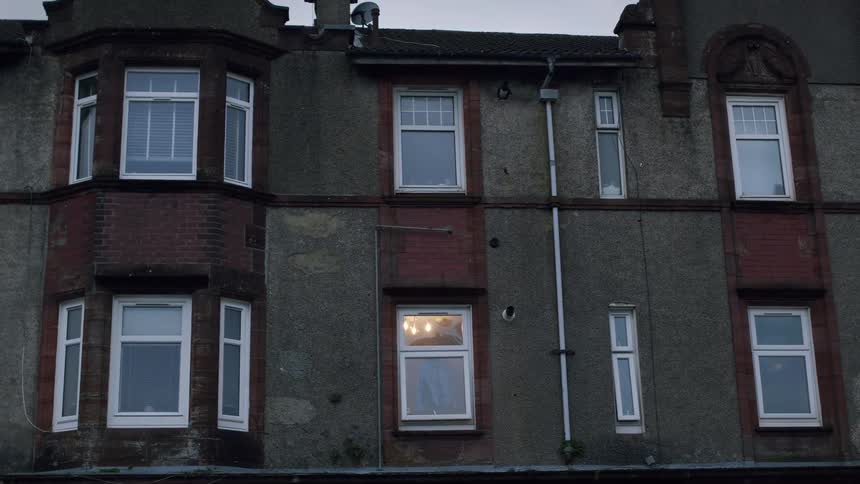 A single light is on in the window of a drab, old apartment building at dusk