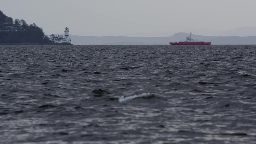 A distant red ship and a coastal lighthouse stand against a grey sky across a choppy, dark sea