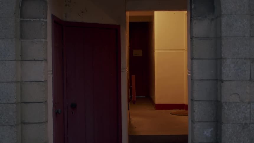 An atmospheric shot of an empty, dimly lit hallway framed by a stone doorway next to a red door
