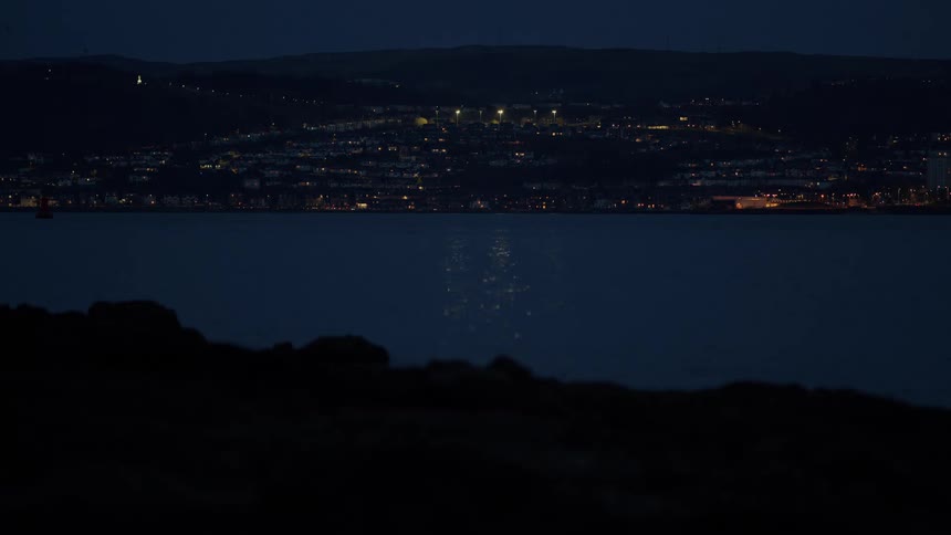 The glowing lights of a hillside town reflect upon the dark, still water at night