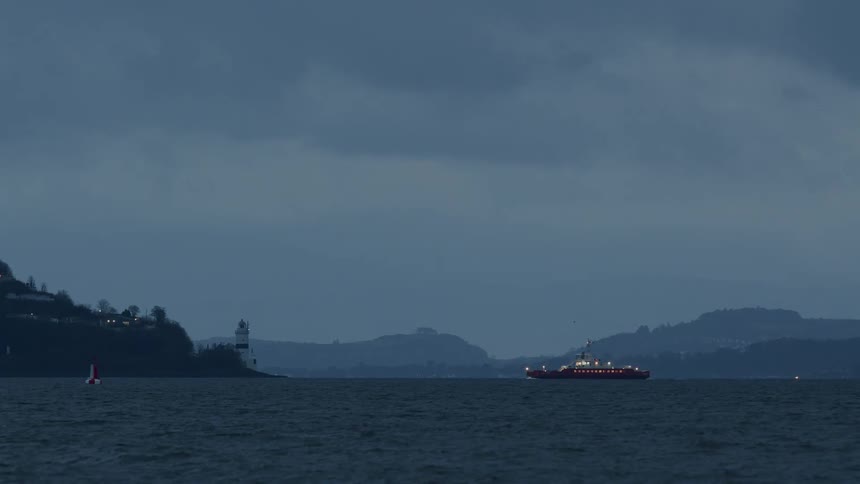 A red ferry with its lights on sails across a bay past a lighthouse on a moody, overcast evening