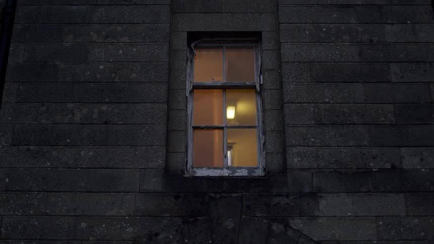 A single illuminated window with a peeling frame set in a dark stone wall at night