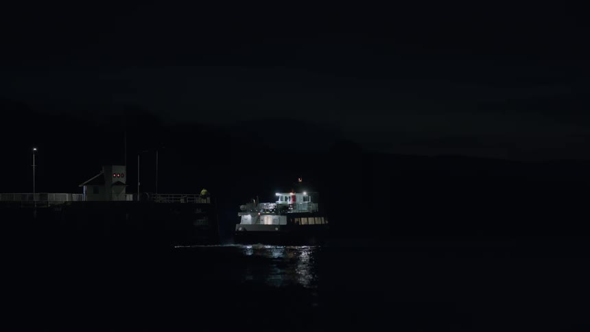A ferry departs from a quiet dock in the dead of night, its lights reflecting on the calm, dark water as it sails away