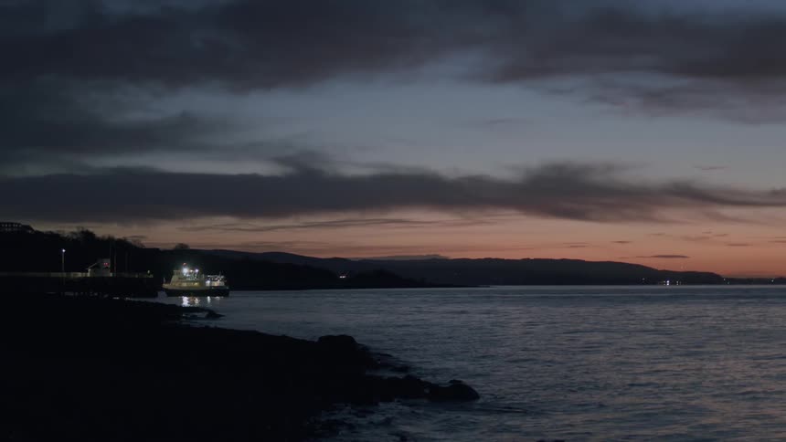 A passenger ferry with its lights on waits at a coastal dock during a beautiful, serene sunset
