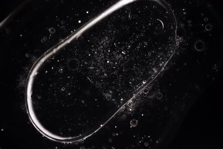 Two clear, particle-filled bubbles slowly merge and combine into a single entity against a stark black background