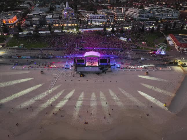 An expansive aerial shot slowly retreats from a lively music festival on the beach, revealing the vast crowd, illuminated stage, and the ocean waves at sunset